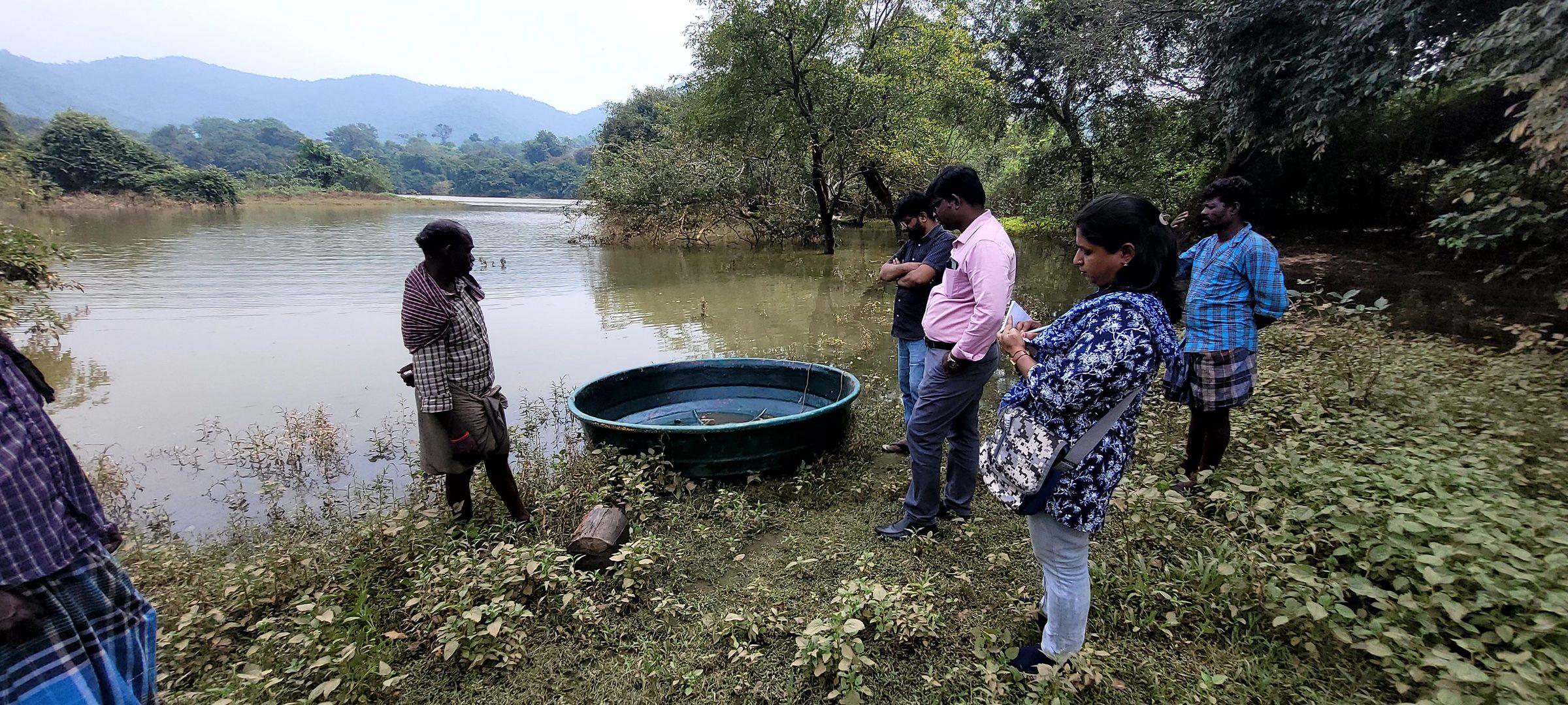 NMCT staff and villagers standing beside a restored water body during a field visit, hills behind them