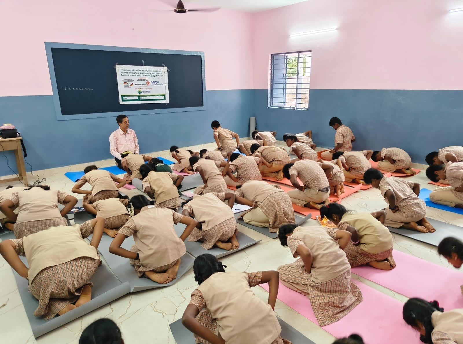 Schoolgirls practising yoga on mats in a classroom, with their teacher