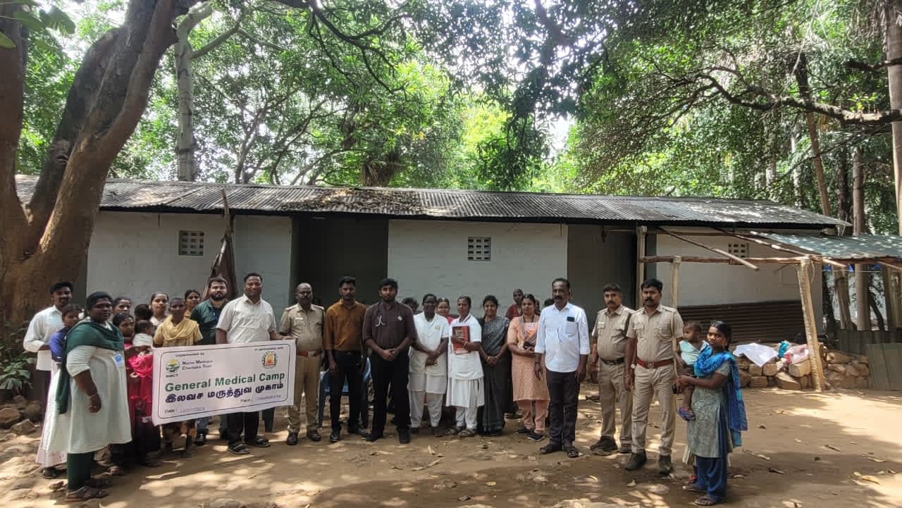 Community members, health workers and forest staff with a General Medical Camp banner outside a village building